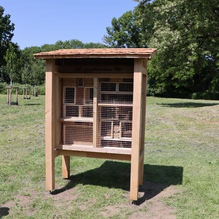 Ein großes Wildbienenhotel aus Holz auf einer Wiese, im Hintergrund sind Bäume