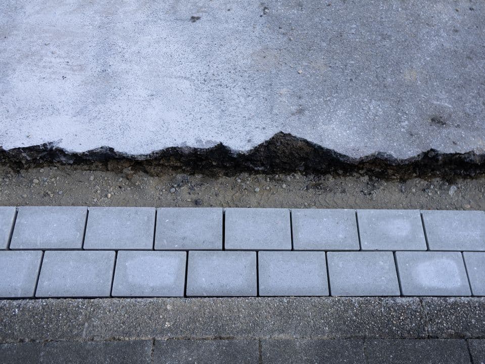 Deckensanierung Man sieht von oben eine Abbruchkante aus grauem Asphalt und graue gepflasterte Steine.